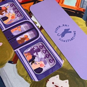 May include: Two illustrated bookmarks. One features a purple train car design with cartoon mice passengers. The other is solid lavender with the text "PAPER_RAT ILLUSTRATIONS" and a rat silhouette. The background is a colorful fabric.