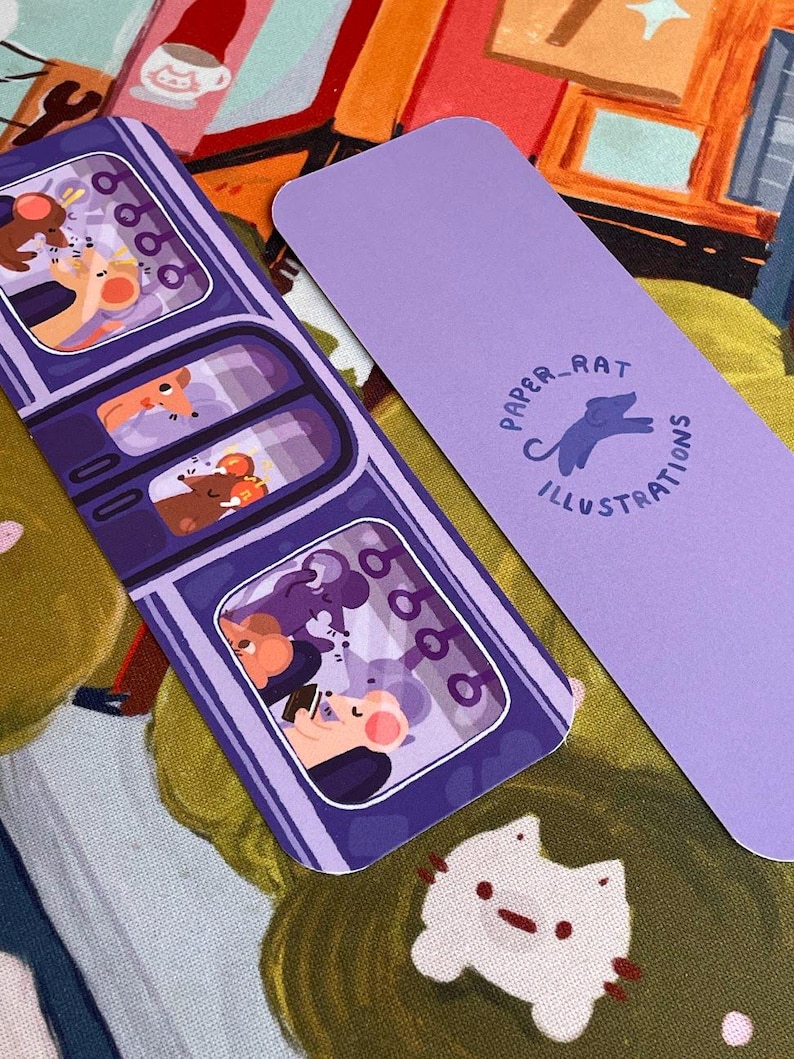 May include: Two illustrated bookmarks. One features a purple train car design with cartoon mice passengers. The other is solid lavender with the text "PAPER_RAT ILLUSTRATIONS" and a rat silhouette. The background is a colorful fabric.