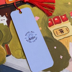 May include: A pale blue bookmark with a red tassel. The bookmark has the text "PAPER RAT & WEIRD SISTERS" and a small illustration. The background shows a colourful illustration with cats and buildings.