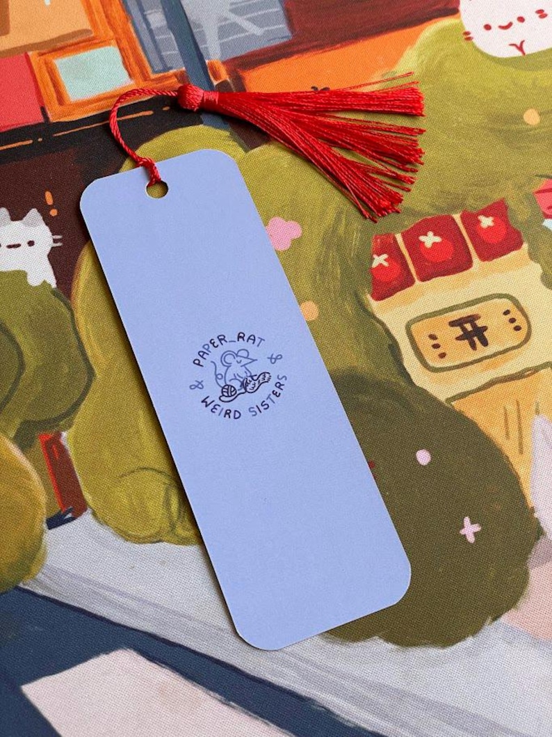 May include: A pale blue bookmark with a red tassel. The bookmark has the text "PAPER RAT & WEIRD SISTERS" and a small illustration. The background shows a colourful illustration with cats and buildings.