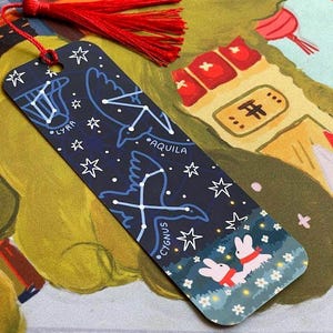 May include: A navy bookmark featuring white constellation designs, including Lyra, Aquila, and Cygnus, with a red tassel. The bookmark has an illustration of two rabbits. The background depicts a starry night scene.