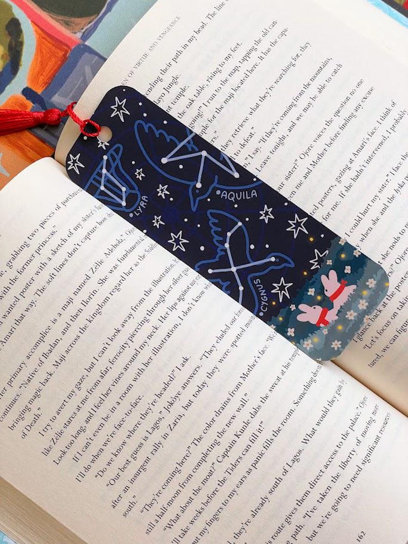 May include: Dark blue bookmark featuring constellations, including Lyra, Aquila, and Cygnus, and white stars. The bottom has a floral pattern and two pink rabbits. A red tassel hangs from the top. The bookmark is in an open book.