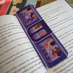 May include: A purple bookmark with three rectangular windows, each featuring a cartoon illustration of two characters. The bookmark is resting on an open book with printed text. The illustrations have a cartoon style.
