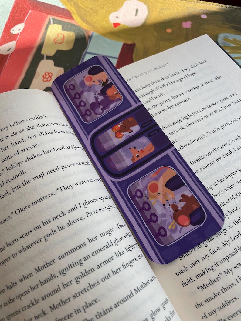 May include: A purple bookmark with three rectangular windows, each featuring a cartoon illustration of two characters. The bookmark is resting on an open book with printed text. The illustrations have a cartoon style.