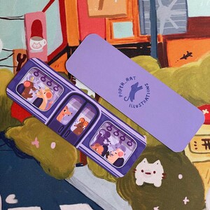 May include: Two purple bookmarks with illustrations. One shows a subway car with cartoon animals, the other has the text "PAPER RAT ILLUSTRATIONS" and a rat silhouette. A small cat-shaped pin and a green object are also visible.
