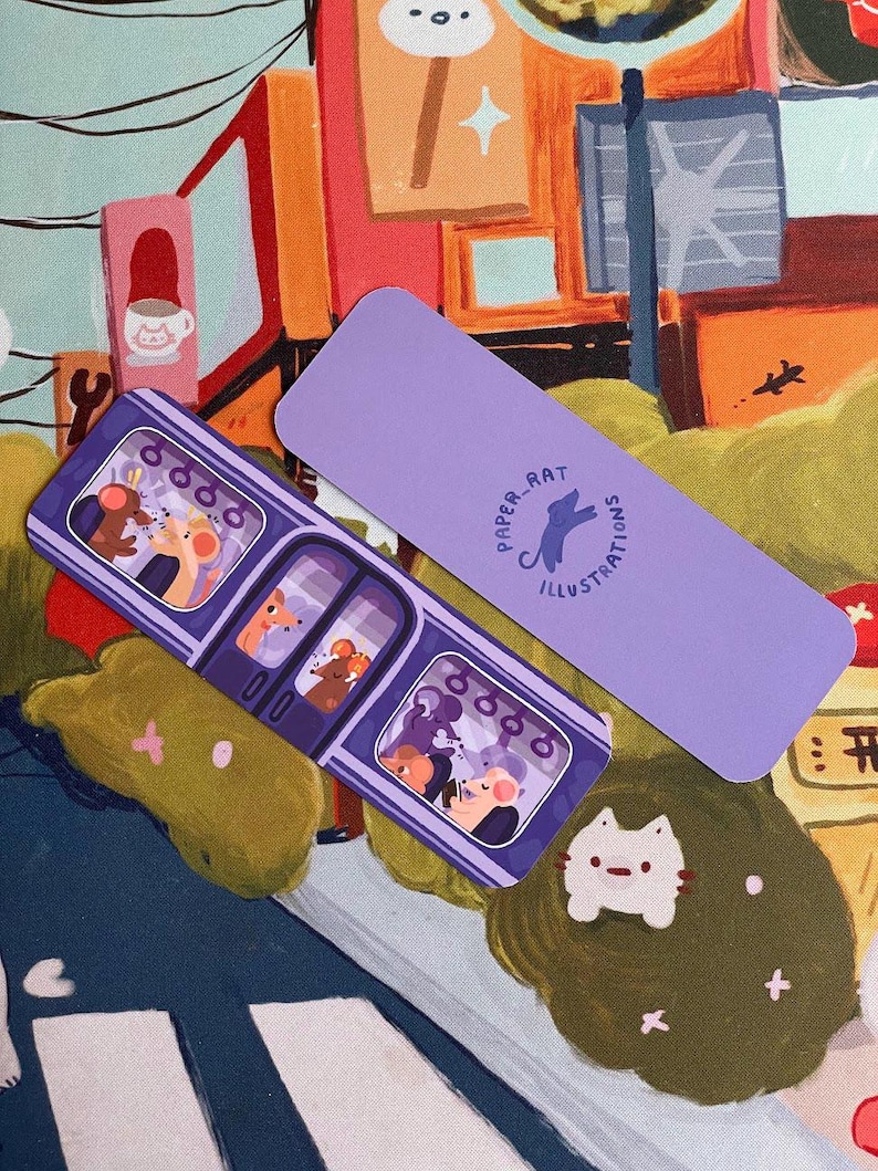 May include: Two purple bookmarks with illustrations. One shows a subway car with cartoon animals, the other has the text "PAPER RAT ILLUSTRATIONS" and a rat silhouette. A small cat-shaped pin and a green object are also visible.