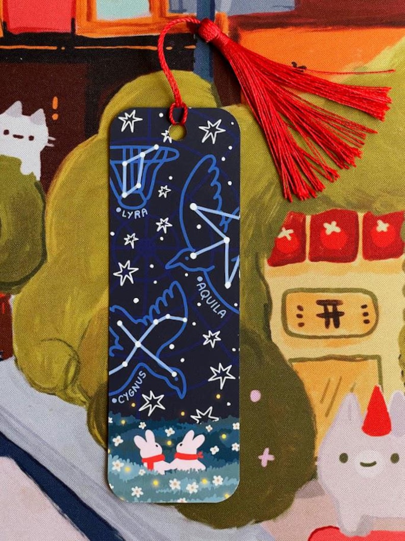 May include: A rectangular bookmark with a dark blue background featuring constellations like Lyra, Aquila, and Cygnus, along with white stars. The bottom of the bookmark shows two rabbits in a field of flowers. A red tassel is attached at the top.