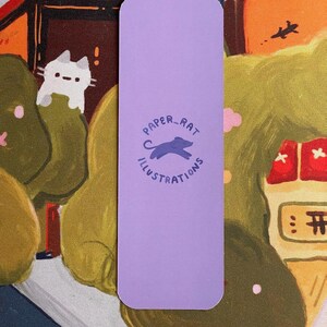 May include: A lavender bookmark with rounded corners featuring the text "PAPER_RAT ILLUSTRATIONS" in a circular design. The illustration includes a silhouette of a rat. The background shows a colorful illustration of a cat and a landscape.
