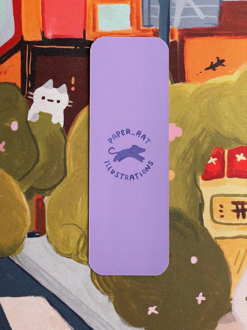 May include: A lavender bookmark with rounded corners featuring the text "PAPER_RAT ILLUSTRATIONS" in a circular design. The illustration includes a silhouette of a rat. The background shows a colorful illustration of a cat and a landscape.