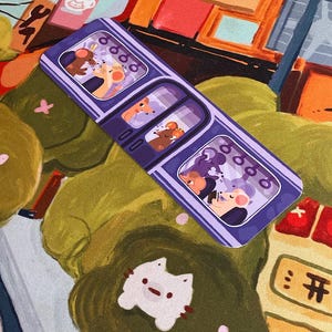 May include: A purple bookmark featuring a train design with three compartments, each showing cartoon mice. The background has a colorful, illustrated scene with a white cat-shaped sticker and other decorative elements. The bookmark is rectangular.