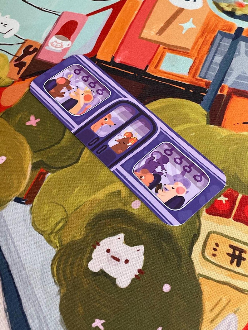 May include: A purple bookmark featuring a train design with three compartments, each showing cartoon mice. The background has a colorful, illustrated scene with a white cat-shaped sticker and other decorative elements. The bookmark is rectangular.