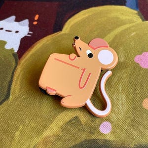 May include: A cartoon-style mouse pin with a light orange body, pink ears, and a white-tipped tail. The pin has a U-shaped design on its belly and is set against a green and yellow patterned background.