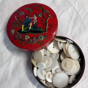 May include: A round red metal tin with a lid featuring a painted illustration of two figures in a landscape with trees and flowers. The tin is filled with a collection of various white, circular, multi-hole buttons of different sizes.