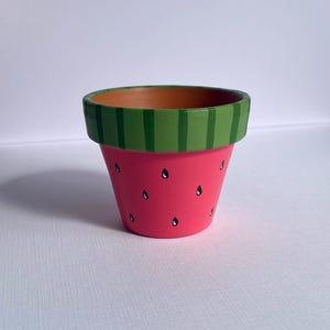 May include: A small, hand-painted terracotta flower pot. The pot is painted to resemble a watermelon, with a pink body and black seed details. The rim is green with vertical stripes.