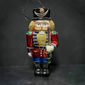Peut inclure: Une figurine de casse-noisette avec un chapeau noir orné d'une étoile et d'une chaîne dorées, une veste rouge à détails bleus et dorés, un pantalon bleu et des bottes noires. La figurine a une barbe dorée, des yeux bleus et tient une épée argentée.