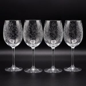 May include: A set of four clear glass wine glasses, each featuring a delicate etched pattern of swirling vines and leaves covering the bowl. The glasses have a slender stem and a circular base, standing upright in a row against a dark background.