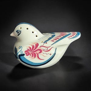 Op de afbeelding: Een crèmekleurige keramische vogelvormige ocarina met een glanzende afwerking. Het instrument is versierd met ingewikkelde blauwe en roze bloemmotieven op de zijkant en vleugels, en bevat verschillende kleine ronde vingergaten aan de bovenzijde.
