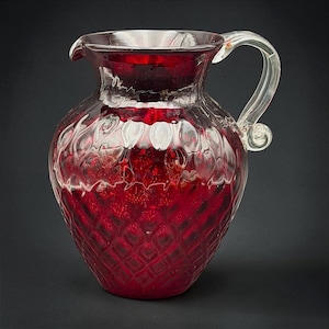 May include: A deep red glass pitcher featuring a textured diamond pattern across the body, a wide circular rim with a small pouring spout, and a clear, curved glass handle attached to the side with a decorative swirl at the base of the handle.