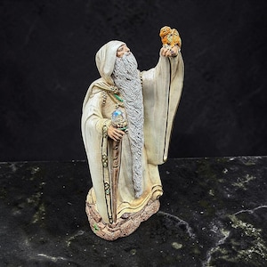 May include: A figurine of a bearded wizard wearing a hooded cream-colored robe, holding a wooden staff topped with a clear iridescent orb in one hand and a small orange dragon perched on the other raised hand, featuring intricate gold trim details.