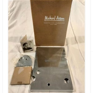 May include: A collection of metal and cardboard components, including a large, rectangular metal piece with circular cutouts, a smaller square cardboard box, and a box with the text "Michael Aram". The items are arranged on a white surface.