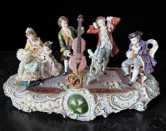 May include: A detailed porcelain figurine depicting a musical ensemble in period attire. The figures, in shades of yellow, green, red, and purple, are arranged on a decorative base with floral accents. A crown is featured in the center.