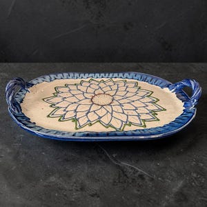 Puede incluir: Una bandeja de servicio ovalada de cerámica con dos asas laterales. El centro presenta un gran diseño floral pintado a mano con pétalos de color crema delineados en azul y verde. El borde tiene un acabado en esmalte azul profundo.