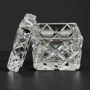 May include: A clear, square glass container with a matching lid, featuring a faceted, diamond-cut geometric pattern across all exterior surfaces. The lid is positioned at an angle, revealing the open interior of the transparent glass box.