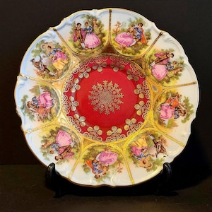 May include: A decorative porcelain plate with a scalloped edge. The plate features a red center with a gold floral design and is surrounded by panels depicting couples in period clothing. Gold trim accents the white and red sections.