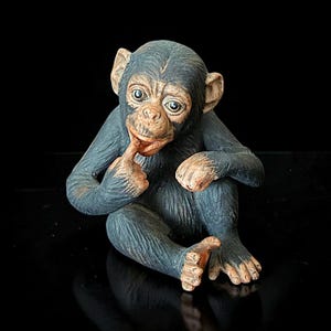 May include: A detailed figurine of a chimpanzee in a seated position, with a finger touching its mouth. The chimpanzee has a dark blue body, tan face, and is set against a black background. The figurine is a decorative item.