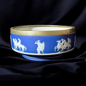 Może przedstawiać: Niebieska ceramiczna miska ze złotą obwódką i białym wnętrzem. Na zewnątrz znajdują się białe płaskorzeźby przedstawiające sceny klasyczne. Miska ma średnicę około 20 cm.
