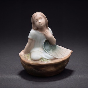 May include: A porcelain figurine of a person with light brown hair wearing a white garment, sitting inside a brown textured walnut shell. The person holds a large, pale green leaf that drapes over their lap, with a smooth, glossy finish throughout.