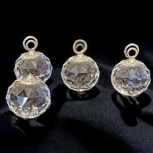 May include: Four clear crystal ball ornaments with silver-colored metal tops and hanging loops. The faceted spheres reflect light, creating a sparkling effect. The ornaments are arranged on a dark, textured surface.