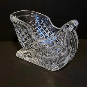 Puede incluir: Un trineo decorativo de cristal transparente con un patrón de diamantes en los lados y un patín curvo. El trineo tiene un diseño detallado y probablemente se utiliza para la decoración navideña.