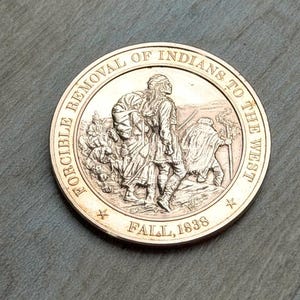 May include: A bronze-colored coin with the text "FORCIBLE REMOVAL OF INDIANS TO THE WEST" and "FALL, 1838" encircling a detailed relief of Native Americans. The coin's design features figures in period clothing, carrying packs and walking.
