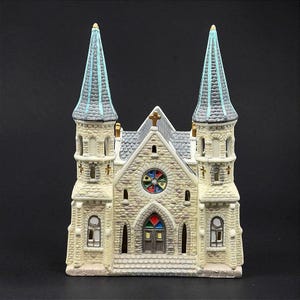 May include: A ceramic miniature church building featuring two tall spires with blue-grey roofs, a stone-textured facade, a central circular stained-glass window with multi-colored panes, and a double-door entrance with a colorful arched transom.