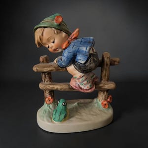 May include: A ceramic figurine of a child wearing a green hat, blue jacket, and patterned socks, leaning over a brown wooden fence. A small green frog sits on the base below the fence, which is decorated with small orange flowers.