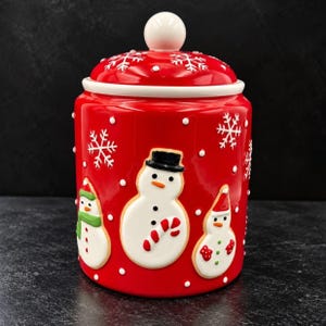 Op de afbeelding: Een cilindrische rode keramische voorraadpot met een bijpassend deksel met een witte knop. De buitenkant is versierd met reliëf witte sneeuwvlokken, kleine witte stippen en drie sneeuwpopfiguren, waarvan er één een zwarte hoed draagt.