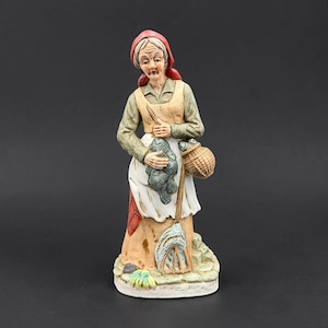 May include: A hand-painted ceramic figurine of an elderly person wearing a red headscarf, a green long-sleeved shirt, and a tan apron. The figure holds a cluster of grey fish in their hands, carries a woven basket, and stands on a stone base.