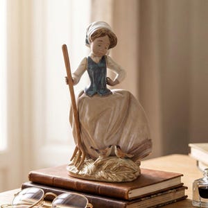 May include: A porcelain figurine of a person wearing a white head covering, a blue vest, and a long beige skirt. The figure holds a tall wooden pitchfork and stands beside two small brown birds perched on a textured base of hay or straw.