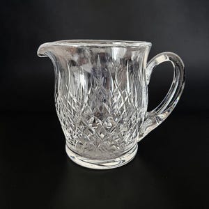 May include: A clear, cut-glass creamer with a handle and a spout. The creamer features a diamond-cut pattern across its body and a rounded base. The handle is curved, and the spout is gently curved for pouring. The creamer is designed for serving.