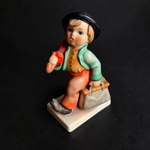 May include: A ceramic figurine of a young person in a black hat, green jacket, orange vest, and orange pants, carrying a red item over their shoulder and a gray suitcase. The figurine is standing on a white base.