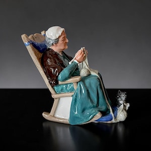 Könnte beinhalten: Keramikfigur einer Frau, die in einem Schaukelstuhl strickt. Die Frau trägt eine weiße Mütze, ein türkisfarbenes Kleid und einen braunen Schal. Ein Wollknäuel und eine Katze liegen zu ihren Füßen. Der Stuhl ist cremefarben mit blauen Akzenten.