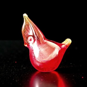 Può includere: Piccola statuetta di uccello in vetro rosso, con becco e coda gialli. L'uccello è in una posa stilizzata con il becco aperto. Il vetro ha una finitura lucida, su sfondo nero.