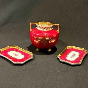 May include: A red and gold decorative ceramic set, including a small vase with handles and two matching trays. The vase has a gold rim and feet. The trays and vase feature a floral design.