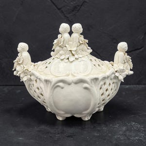 Puede incluir: Un cuenco de cerámica color crema con tapa, que presenta un intrincado patrón de celosía y figuras de querubines esculpidas adornadas con detalles florales en la tapa y los lados. El cuenco tiene un acabado liso y brillante y se apoya en pies.