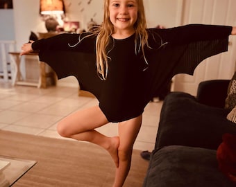 May include: A black long-sleeved leotard featuring integrated bat-wing style fabric panels that extend from the sleeves to the torso, creating a distinct wing-like silhouette when the arms are outstretched, made of a solid black fabric.