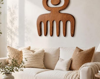 May include: A large, dark brown wooden wall art piece shaped like a traditional African comb with a rounded top and five vertical tines. The object is mounted on a plain, light-colored wall above a sofa, featuring a smooth, polished wood grain finish.