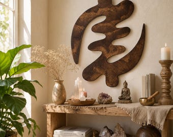 May include: A dark brown wooden Adinkra symbol wall art piece, mounted on a light-coloured wall. Below, a rustic wooden table displays a small Buddha statue, crystals, and candles. The table is part of a room with a window and a large potted plant.