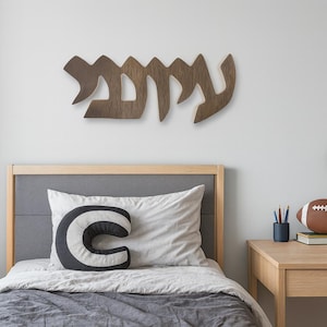 Puede incluir: Un adorno de pared de madera con escritura hebrea está montado sobre una cama. Una almohada decorativa con forma de la letra "C" descansa sobre la cama. Un balón de fútbol americano y una pequeña planta en maceta están sobre un escritorio de madera.