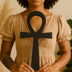 May include: A black ankh symbol, a cross with a loop at the top, is held in front of a person. The ankh is a symbol of life and is often used in spiritual practices. The person is wearing a tan dress.